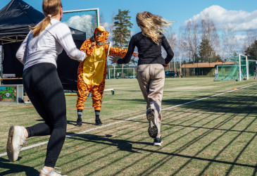 Kaksi nuorta juoksee urheilukentällä ja antaa läpsyn tiikeripukuiselle henkilölle. Kuva: Essi Kultanen.