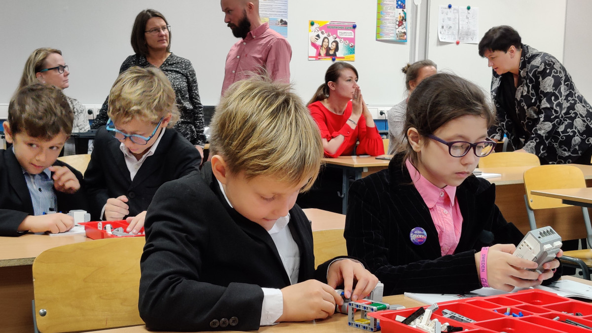 Oppilaat rakentavat pulpeteissa legorobotteja. Henkilökuntaa taustalla.