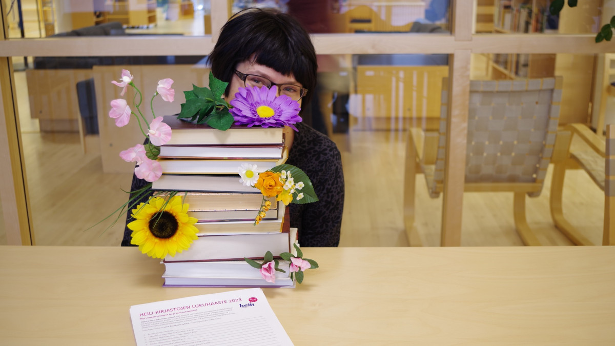 A woman peeks out from behind a stack of books, in the foreground is a reading challenge form.