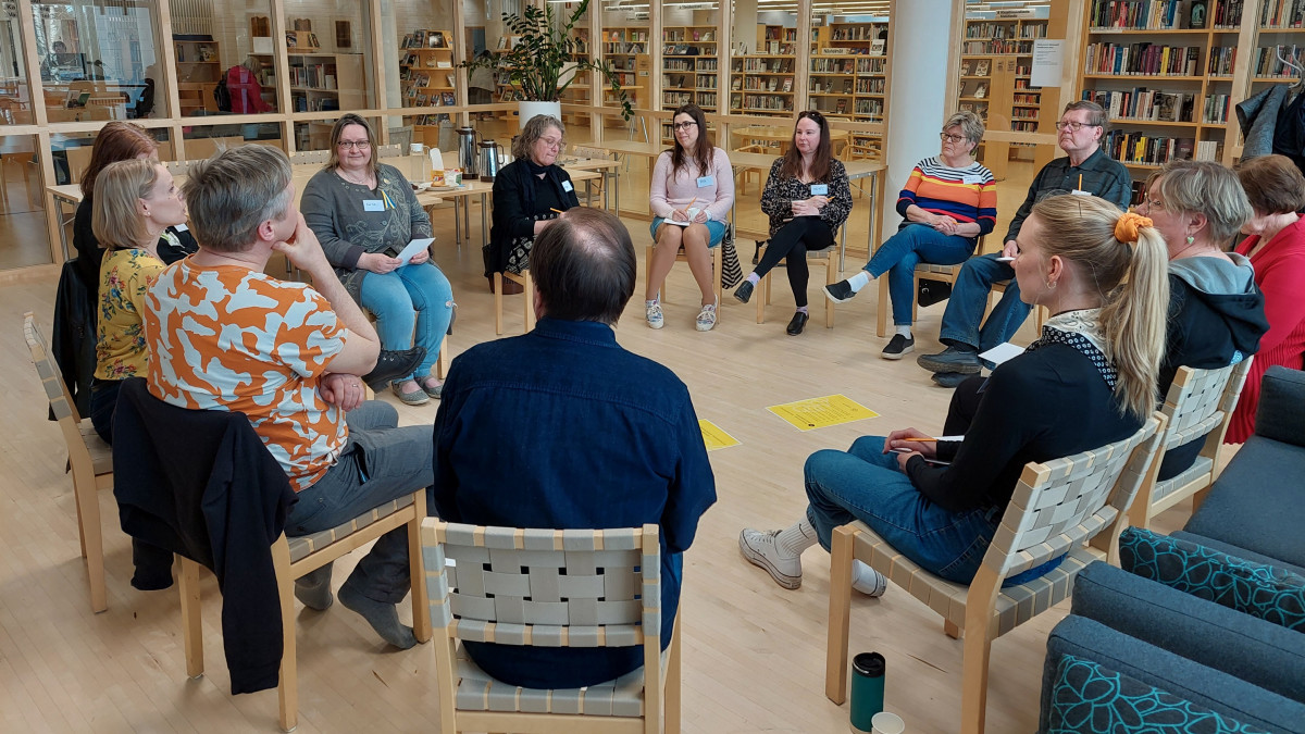 13 people sitting in a circle, the library shelves and customers can be seen in the background.