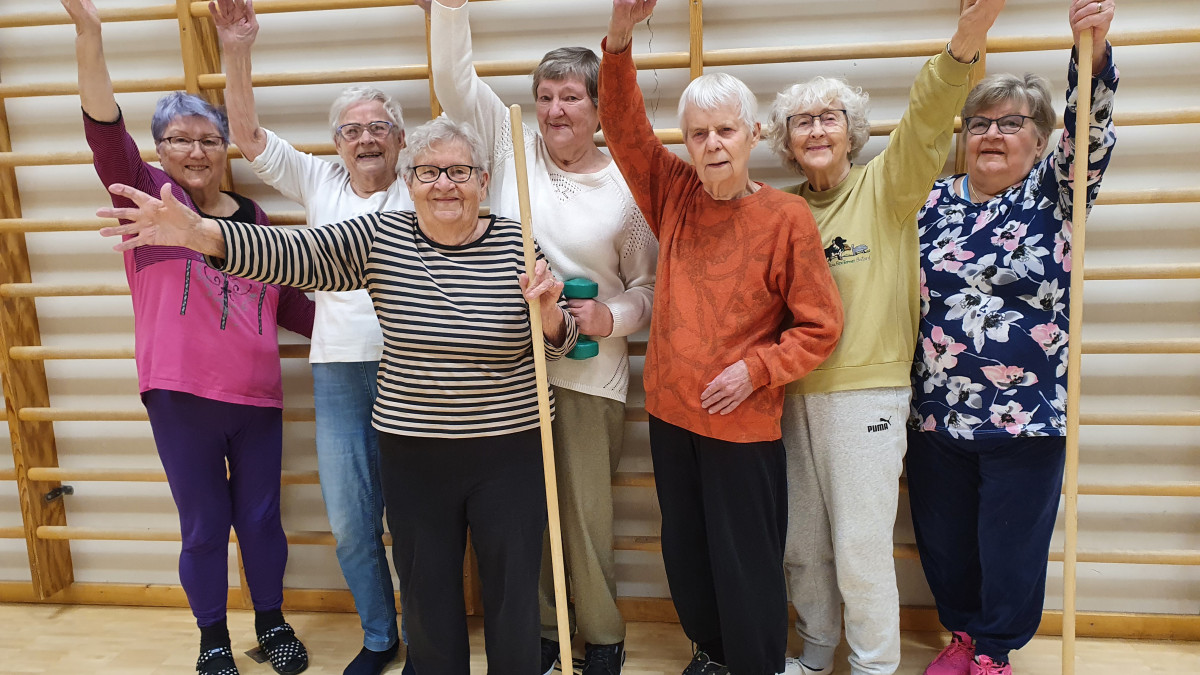 Voimaa vanhuuteen -ryhmän seitsemän naista nostaa käden ilmaan puolapuiden edessä.