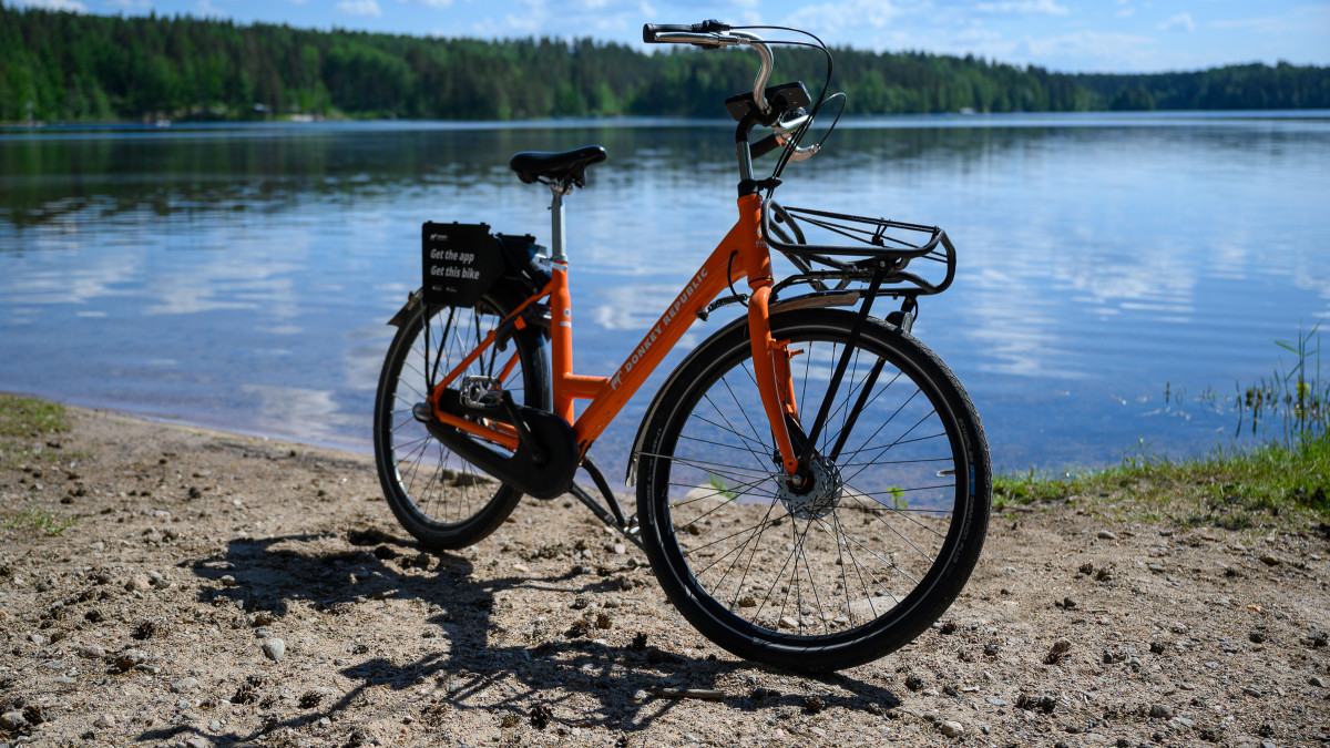 Оранжевый городской велосипед на песчаном пляже, на фоне озера, леса и голубого неба.