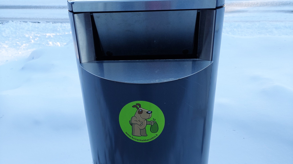 A green outdoor trash can with a dog poop trash sticker glued to it.