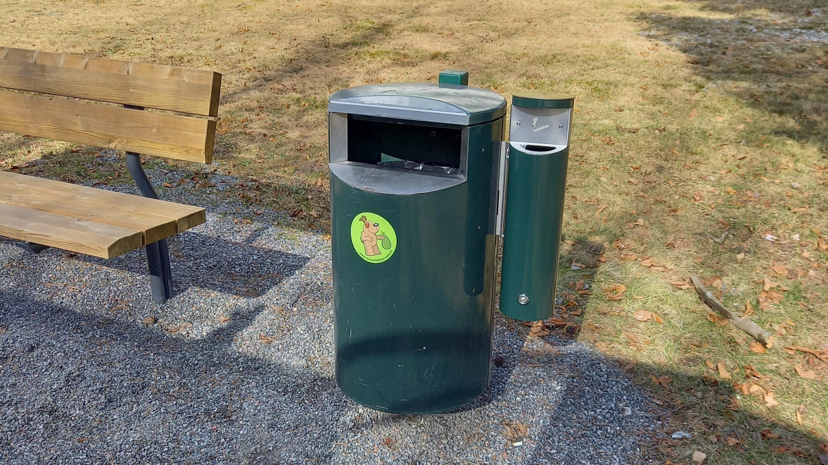 Зеленый мусор рядом со скамейкой в ​​парке. Сбоку от мусорного бака установлен мусорный контейнер.