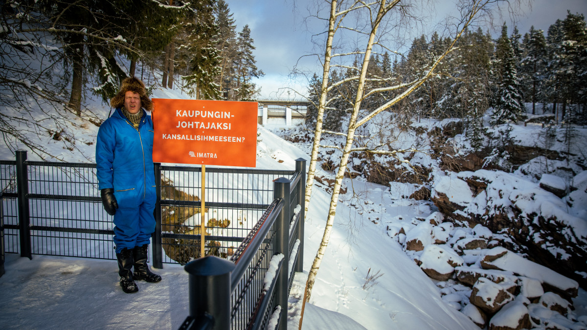 A man in a fur hat stands on the shore of Imatrankoski. The text on the sign: to be the mayor of the national wonder.