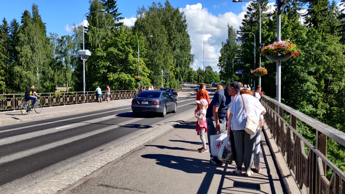 Cars drive on the Imatrankoskentie bridge. There are cyclists and pedestrians on light traffic lanes. Sun shines.