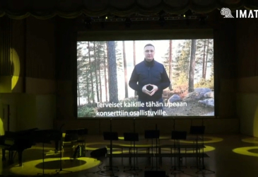 A man on the big screen on the stage of the concert hall, On the screen the text "Greetings to everyone attending this wonderful concert."