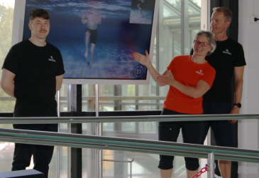 Fitness instructors stand at the edge of the pool and point to the screen.