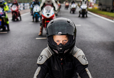 The moped boy looks into the camera at the racing motorcyclists behind him at the starting line.