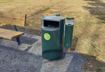 Green trash next to the park bench. A dustbin has been installed on the side of the bin.
