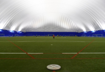 The indoor gym has a green artificial grass field and a white roof.