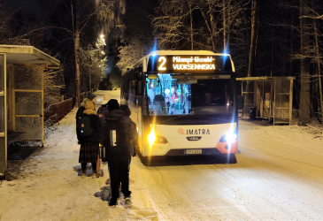 Linja-auto saapumassa pysäkille ja koululaiset odottamassa sitä talvisäässä.