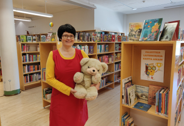 Kirjastonhoitaja Tiina Rossi seisoo lasten kirjaosastolla nalle sylissä.