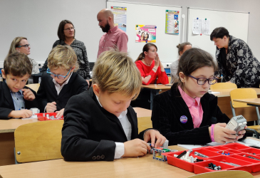 Oppilaat rakentavat pulpeteissa legorobotteja. Henkilökuntaa taustalla.