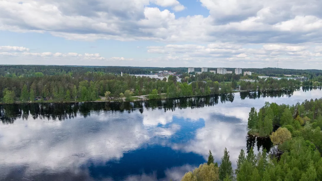 A landscape picture with the calm surface of Vuoksi in the foreground and the towering towers of Mansikkala in the background.